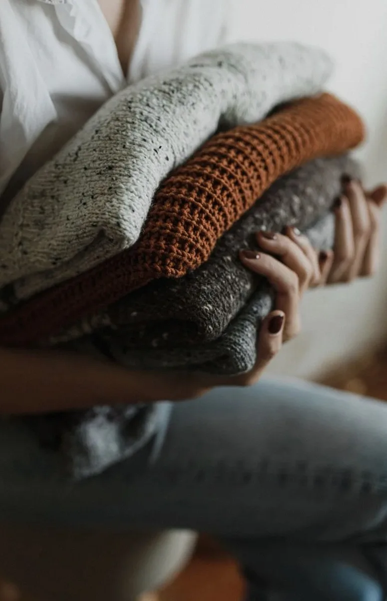 Hands holding a stack of folded knitwear in grey, cream and rust