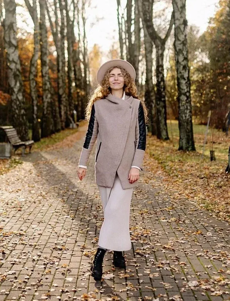 A woman in a long coat and hat standing on a leaf-covered path