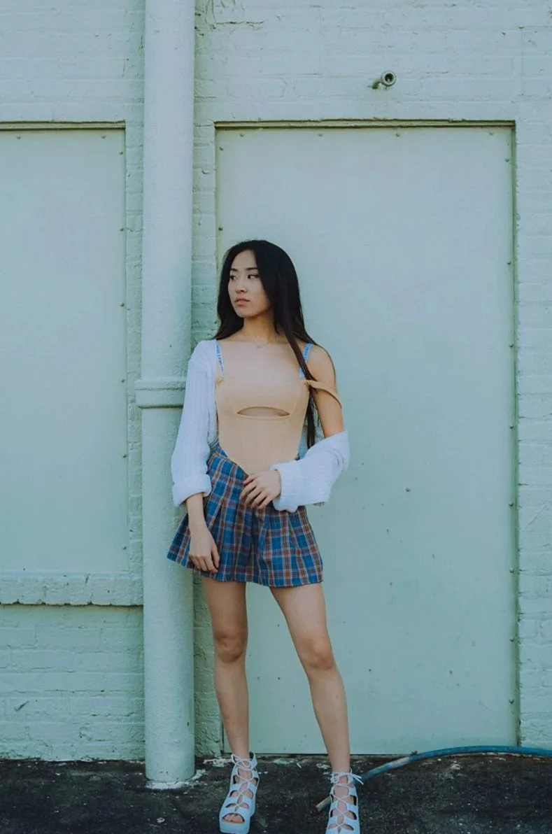 Woman standing against a pale door in a plaid skirt and white cardigan