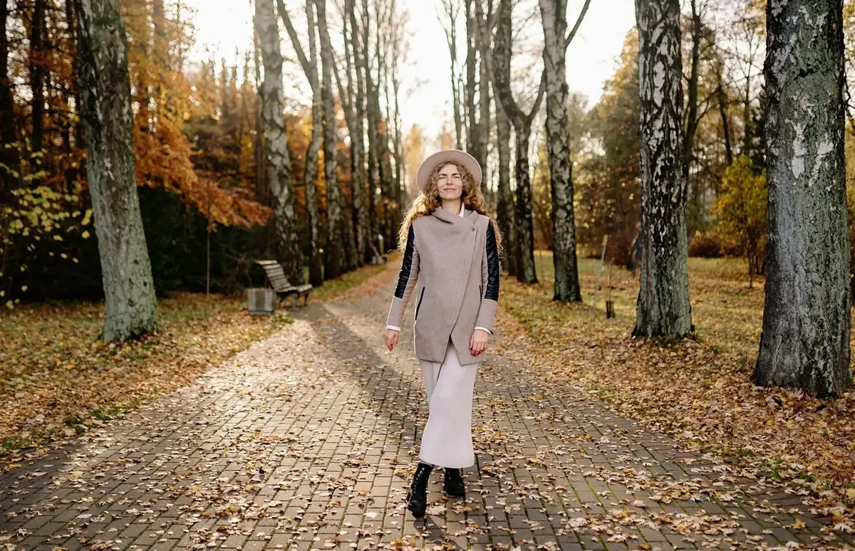A woman in a grey waistcoat and wide-brimmed hat walking down an autumn tree-lined path